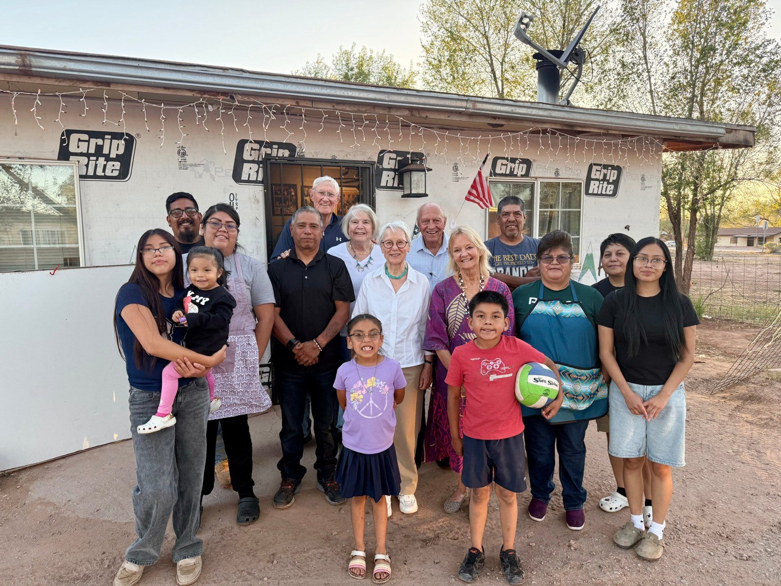 A large group of family and friends of all ages standing together outside a house at dusk, with string lights along the roofline